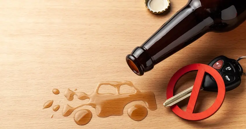A top-down flat lay arranged on a light natural wood surface features an overturned dark brown glass beer bottle lying on its side with its silver bottle cap removed and placed separately at the top of the frame- a large puddle of spilled amber beer that has spread across the wood surface in the rough shape of a vintage automobile with scattered droplets surrounding it and a black electronic car key fob with multiple buttons on a metal keyring positioned beside a three-dimensional red prohibition circle slash symbol โ a cleverly composed conceptual image delivering a clear anti-drunk driving message by connecting alcohol consumption directly to the keys and vehicle that can lead to a deadly drunk driving accident and could use help from Texas Lawyers 365.