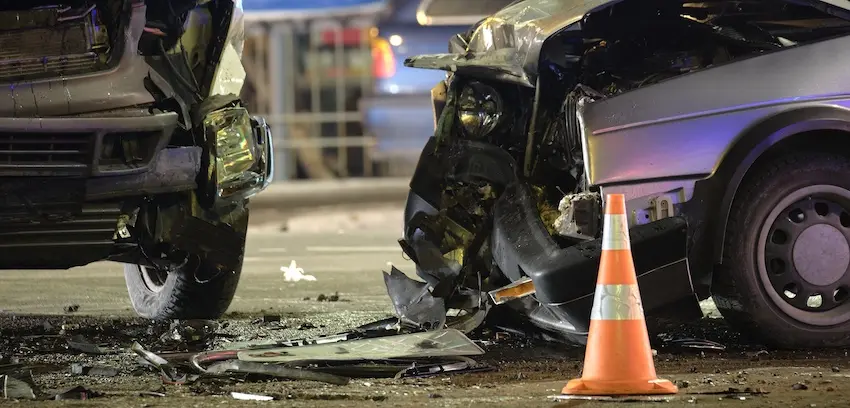 A nighttime urban collision scene photographed- in Texas- at ground level shows two vehicles with catastrophically destroyed front ends locked together at the point of impact- the closer vehicle's entire hood- grille- and headlight assembly reduced to a tangled mass of crumpled metal- shattered plastic- and torn wiring scattered across the wet pavement- while a bright orange and silver reflective traffic safety cone stands upright on the road beside the wreckage and blue and yellow artificial lighting from nearby structures casts a cold glow across the scene โ depicting the catastrophic level of vehicle destruction commonly associated with a high-speed drunk driving accident on a city street after dark.