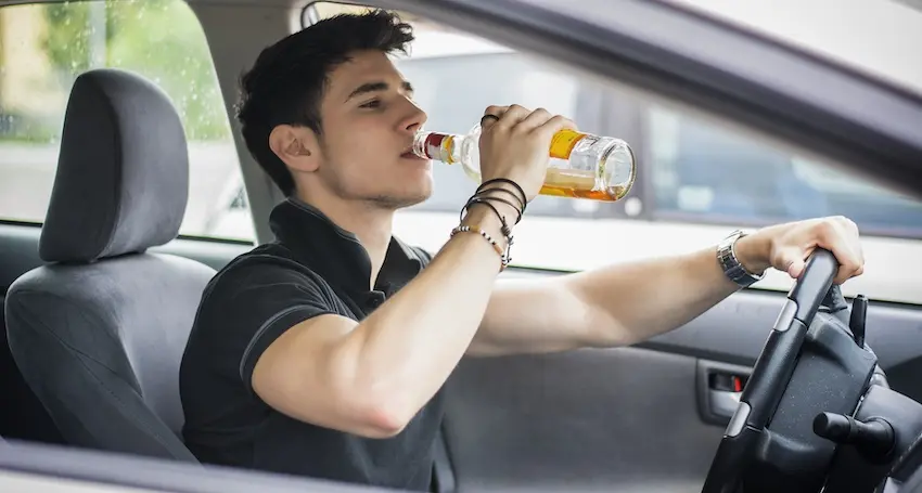 A young male driver with short dark hair- beaded bracelets- and a wristwatch wearing a black polo shirt sits behind the steering wheel of a vehicle with one hand gripping the wheel and the other hand tipping a clear glass bottle filled with amber-colored beer directly to his lips while actively driving with a blurred gray headrest- passenger window- and sunlit exterior visible in the background โ a direct and dangerous depiction of driving under the influence that illustrates the reckless behavior responsible for the majority of drunk driving accident fatalities and injuries in Texas.