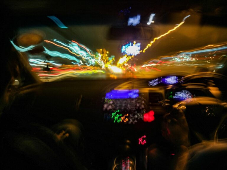 A long-exposure photograph taken from inside a moving vehicle at night captures the driver's silhouette on the left side in near darkness while the entire windshield view is consumed by a dizzying smear of wildly distorted city lights โ streaking ribbons of teal- yellow- orange- red- green- and blue neon and streetlight trails blur chaotically across the frame in every direction with the illuminated dashboard instrument cluster glowing in purple and green tones beneath the rearview mirror โ visually simulating the dangerously impaired and disoriented perspective of a driver operating a vehicle while intoxicated- the kind of impairment that directly causes a drunk driving accident and could use help from Texas Lawyers 365.